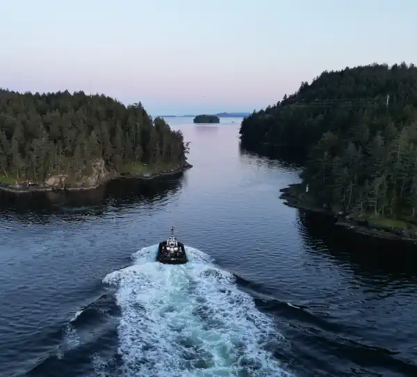 Aerial photo showing a tugboat approaching a narrows between two land masses; a small island is visible in the distance and the sky and water are dusky purple