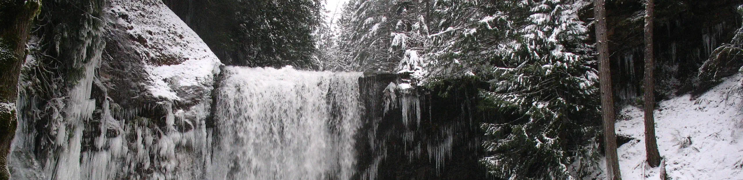 Photo showing the uppermost part of a wide waterfall in winter, surrounded by icicles.