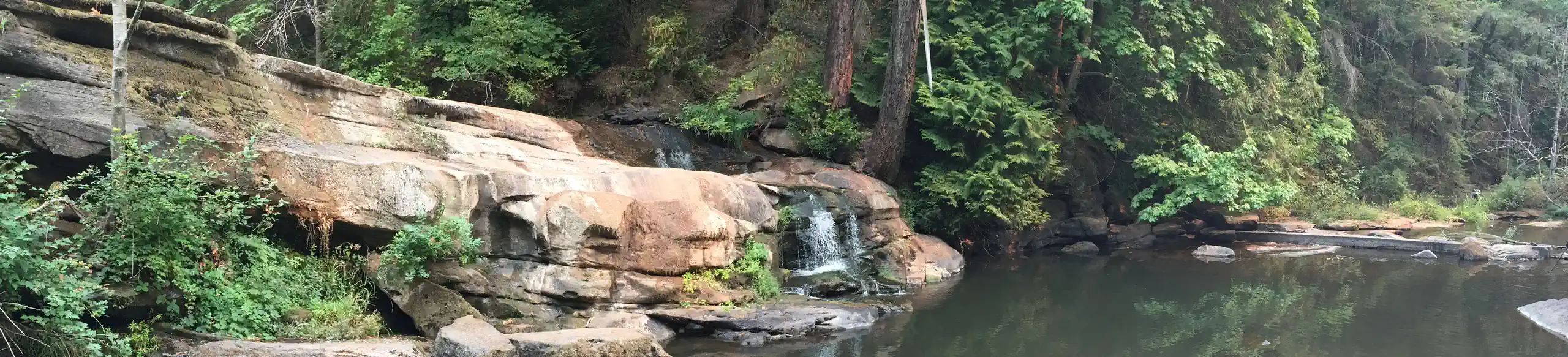 Photo showing a mostly-dry waterfall with a dark pool below - layers of rock form a long wall about 10 feet tall over which the low summer creek leaves an algae stain