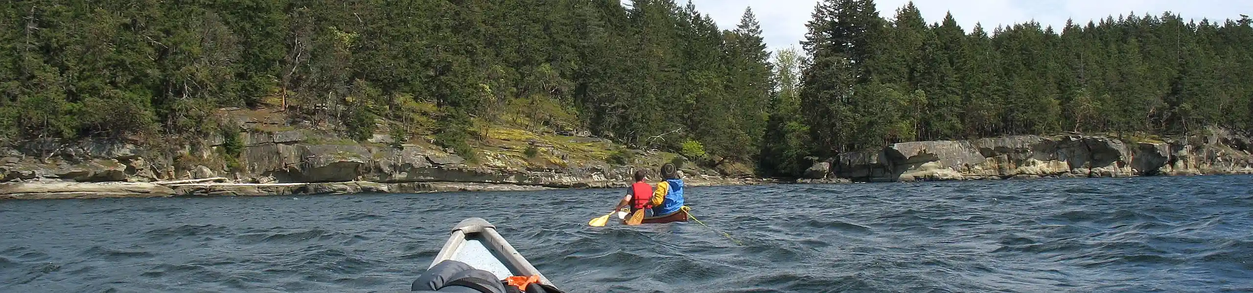 Photo showing two men paddling a canoe in choppy waters toward a nearby shoreline of steep sandstone banks characteristic of BC's Southern Gulf Islands