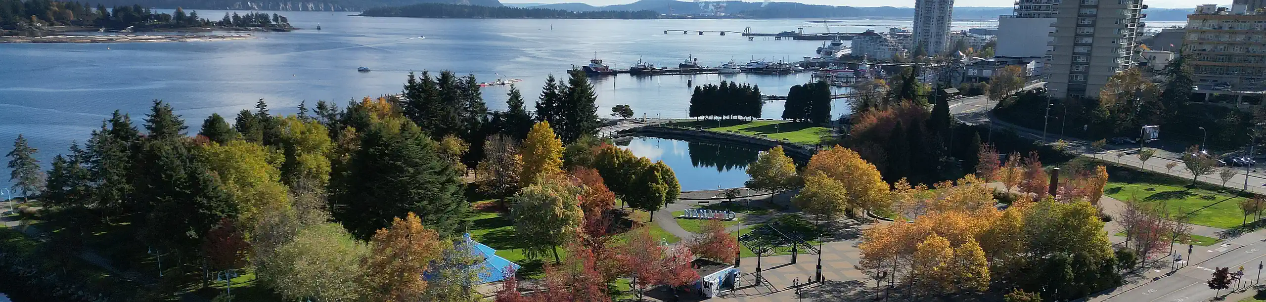 Aerial photo looking down at Nanaimo Harbour with Maffeo Sutton Park in the foreground, an autumnal scene with contrasting shadows and bright orange foliage framing the city park.