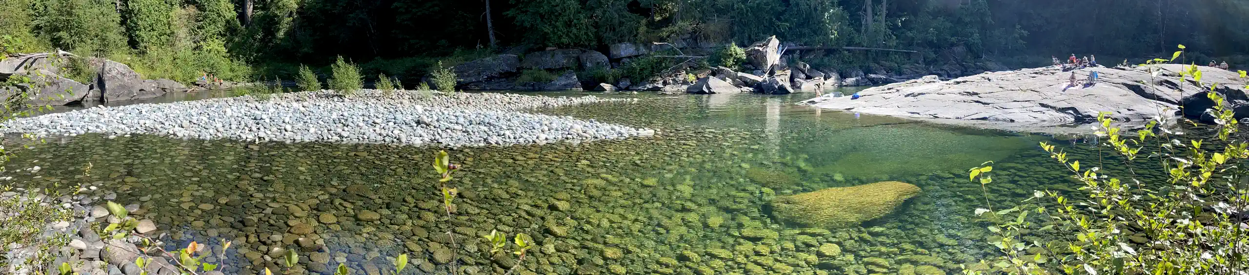 Photo showing a swimming hole at Nanaimo River with a large rock on which some bathers are seated, while an inviting clear pool shows in the foreground.