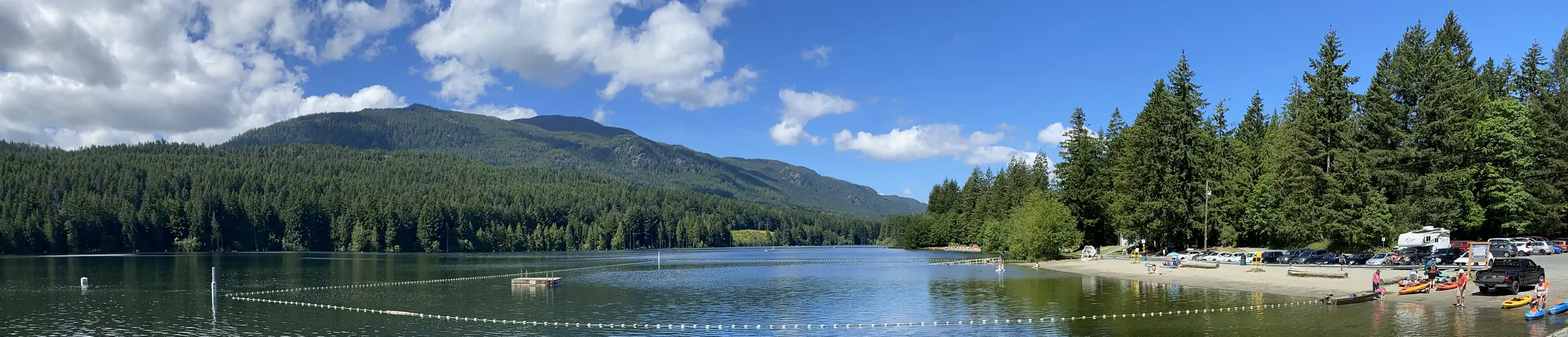 Photo looking at a pretty lake backed by a mountain and forests, with a recreational beach and swimming area in the foreground.